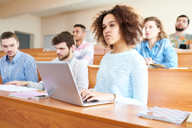 Pensive concentrated young multiethnic students sitting at university desk and attentively listening to lecturer, serious African-American girl with Afro hairstyle typing on laptop