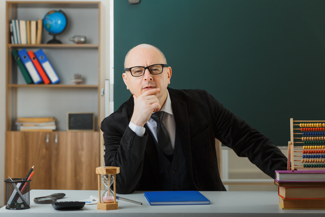 man teacher wearing glasses checking class register looking at camera with confident smile sitting at school desk in front of blackboard in classroom