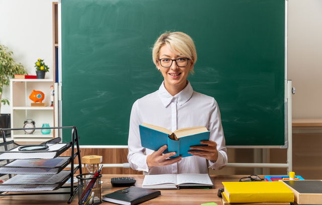 smiling young blonde female teacher wearing glasses sitting at desk with school tools in classroom holding open book looking at camera
