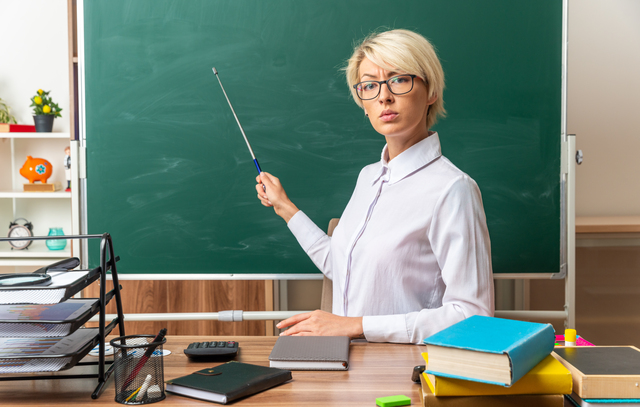 strict young blonde female teacher wearing glasses sitting at desk with school tools in classroom pointing at chalkboard with pointer stick looking at camera